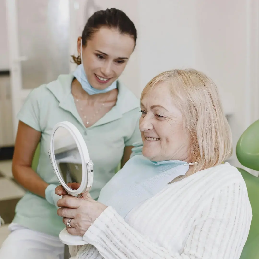 An older patient smiling into a hand mirror in the dental chair while a team member looks on
