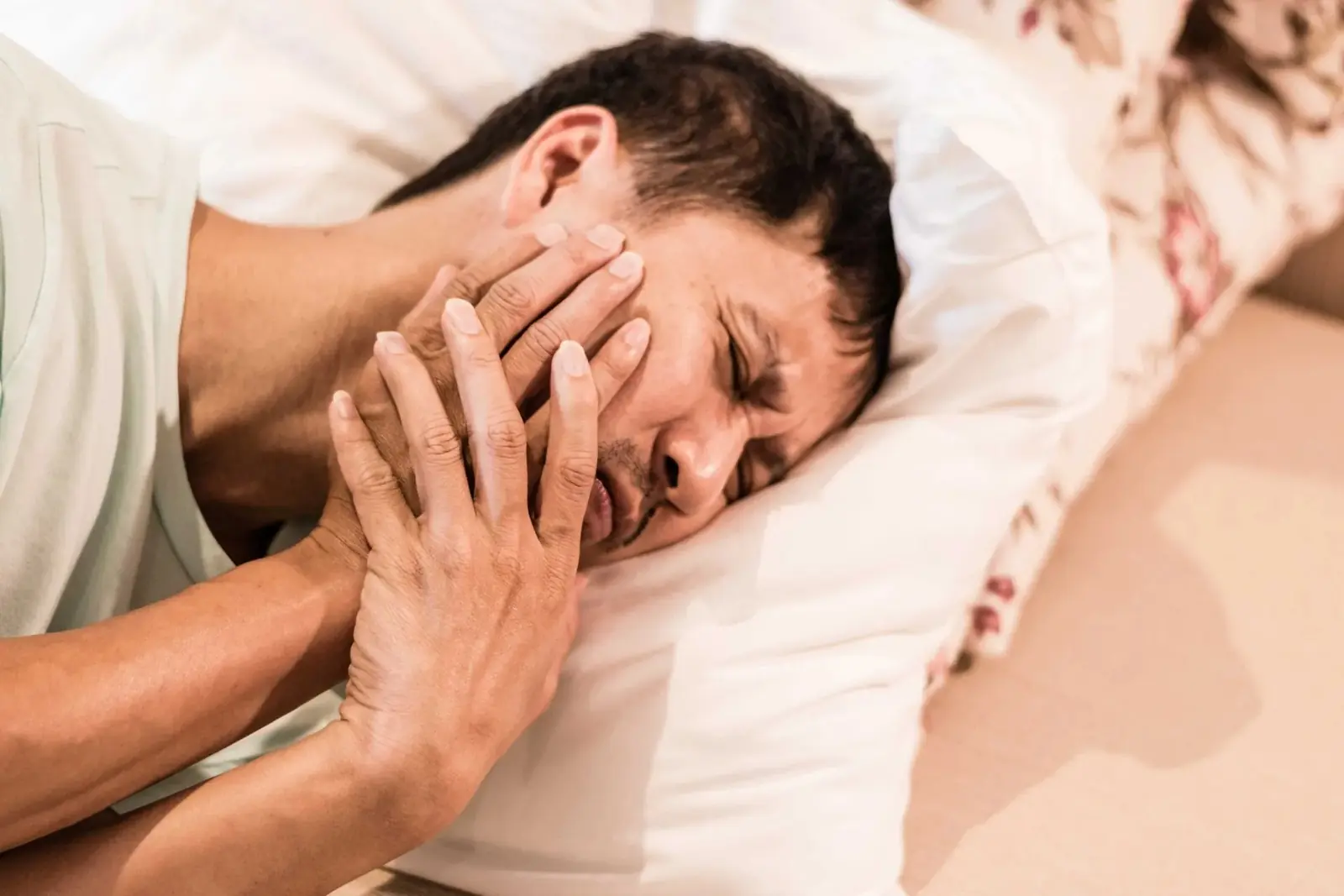 A person lying awake in bed, eyes shut, holding a hand against their jaw